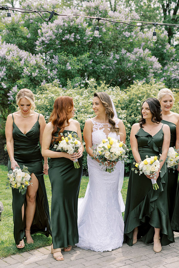 A bride and her bridesmaids assemble for a group photo in a beautiful garden setting. Bridesmaids are wearing forrest green dresses and peal drop earrings.