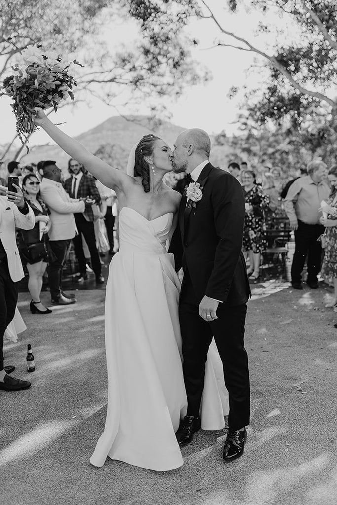 An elated bride and groom share a passionate kiss as they walk down the aisle. Groom is wearing a custom black suit and pink rose. Bride is wearing a strapless white gown, veil and pearl bridal earrings.